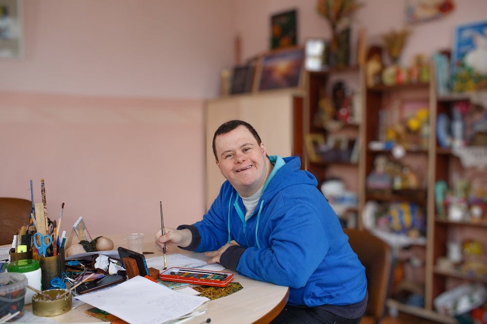 Man with Down syndrome happily engaging in creative work at a cluttered table filled with various art supplies, in a room with shelves of colorful items.