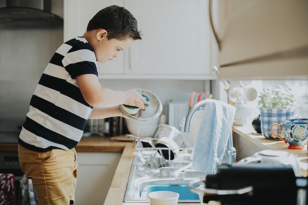 Young man with Down syndrome focused on washing a dish at a kitchen sink, with a running faucet and kitchen items around.