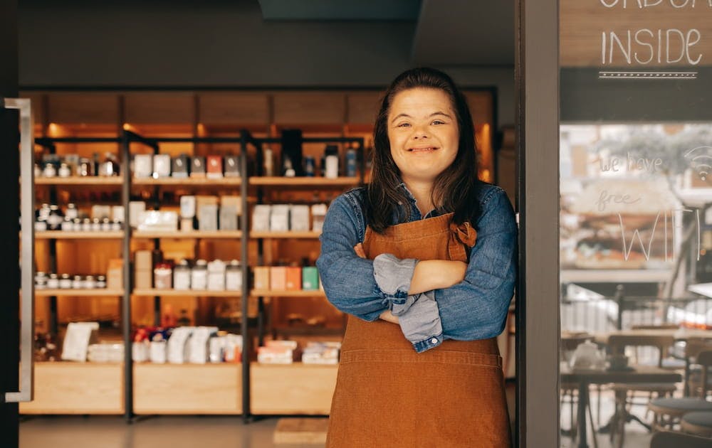 A cheerful woman with Down syndrome wearing an apron, standing with crossed arms in front of a coffee shop interior with shelves of products.